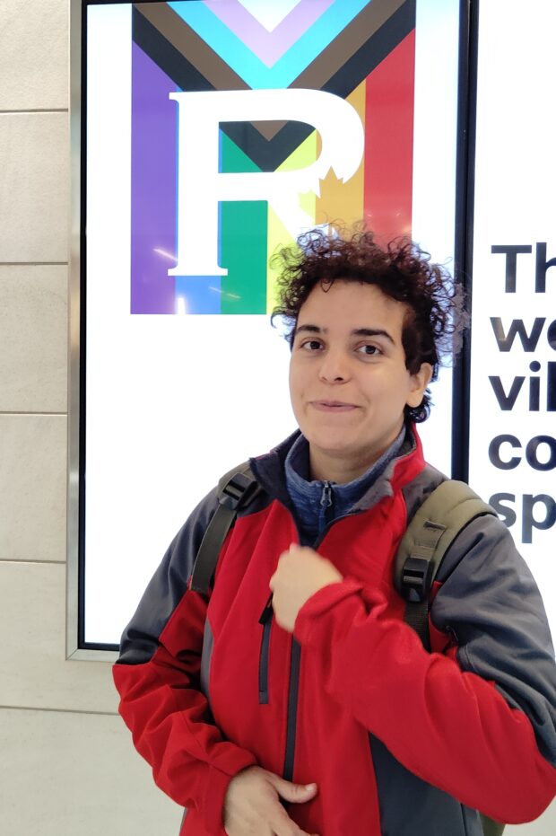 Person standing indoors wearing a red and gray jacket in front of a Pride-themed display.