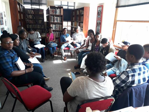 Group seated in a circle in a library room, holding papers during a meeting.