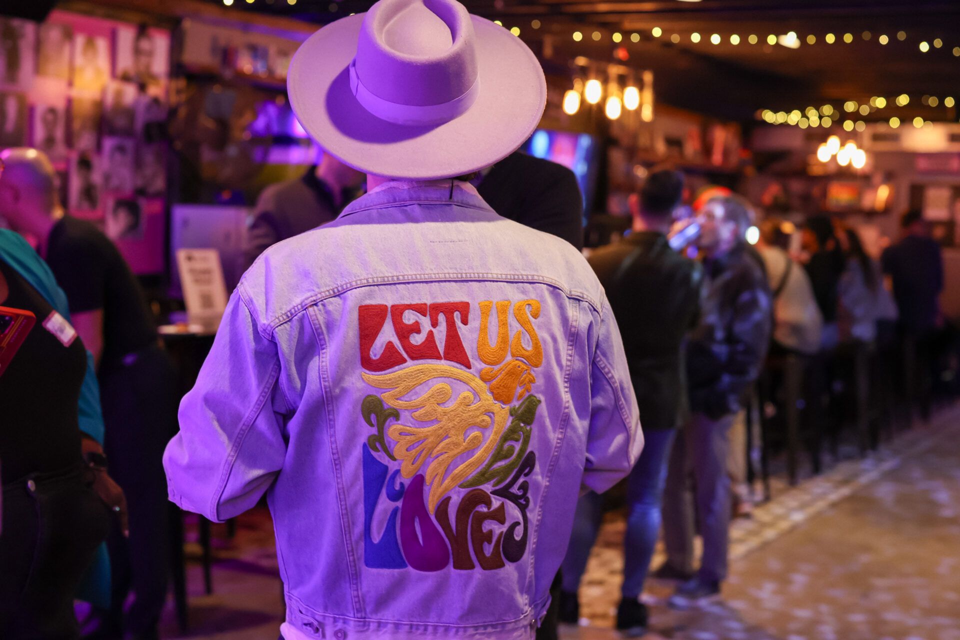 Person wearing a lavender hat and denim jacket with colourful text reading “LET US LOVE,” standing in a warmly lit, festive bar setting.