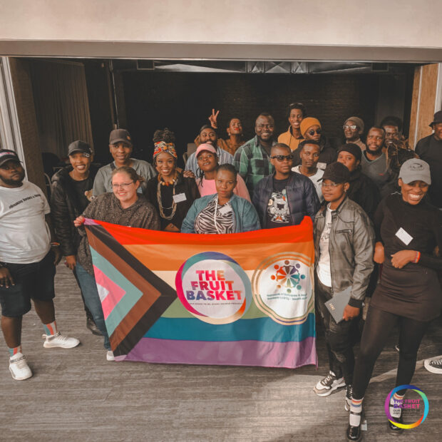 Group photo indoors holding a Pride flag that reads “THE FRUIT BASKET.”