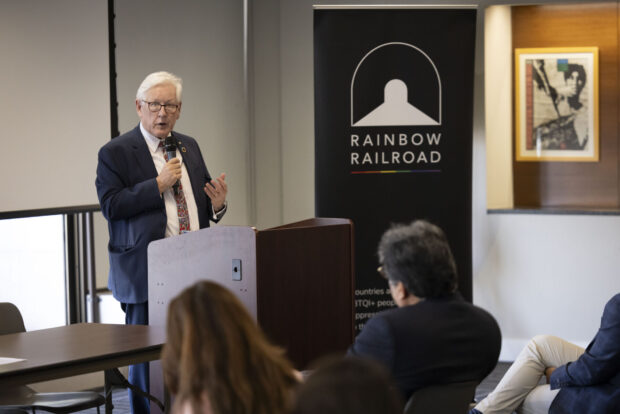 A speaker addresses an audience at a Rainbow Railroad event beside a branded banner.