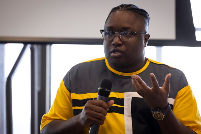 A person in a yellow and gray shirt speaks into a microphone during an event.