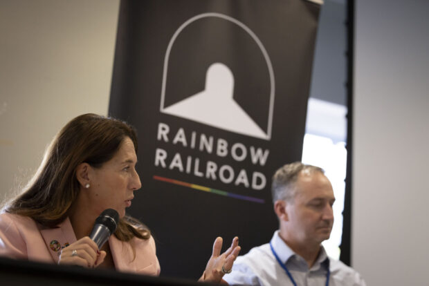 Two panelists speak beside a Rainbow Railroad banner at a formal event.