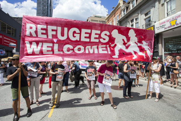 People holds a large red banner reading “REFUGEES WELCOME!” during a street march.