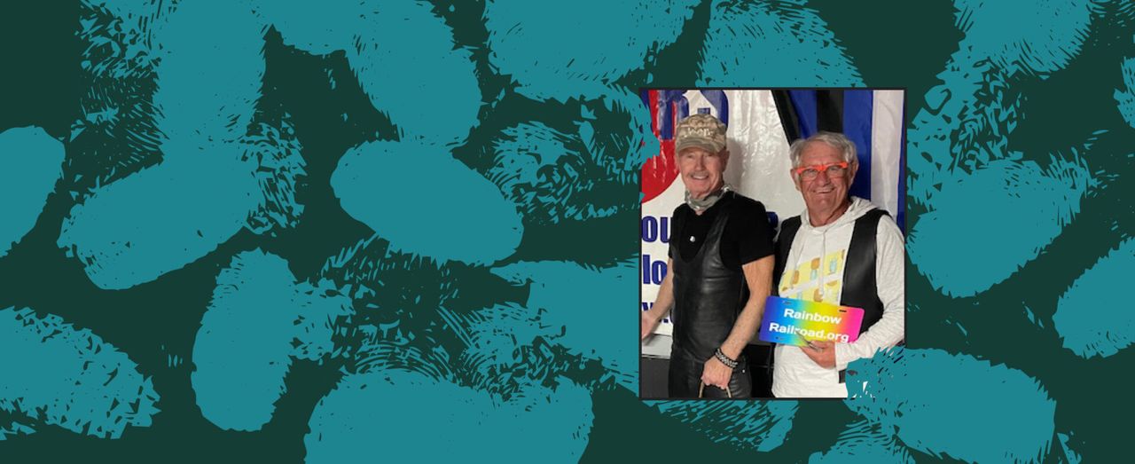 Two people smiling together at an event holding a Rainbow Railroad sign, with a patterned teal background.