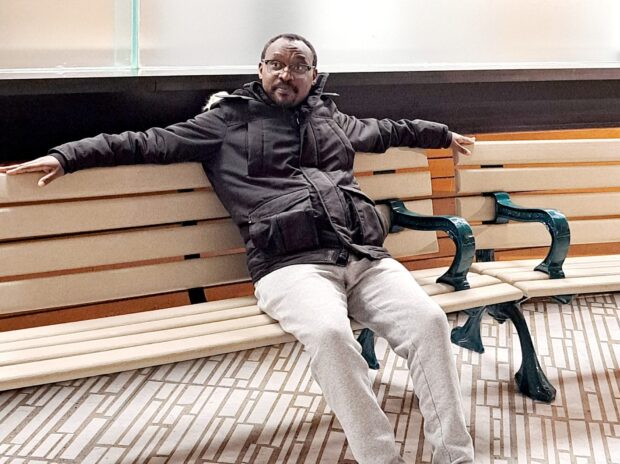 Person in a dark jacket and light pants sitting on a wooden bench.