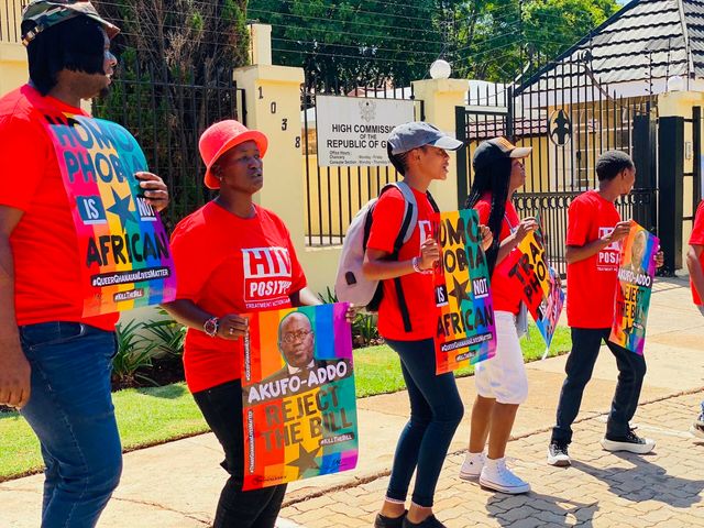 Protesters in red shirts outside a government building holding signs reading “Homophobia is not African” and “Reject the bill.”