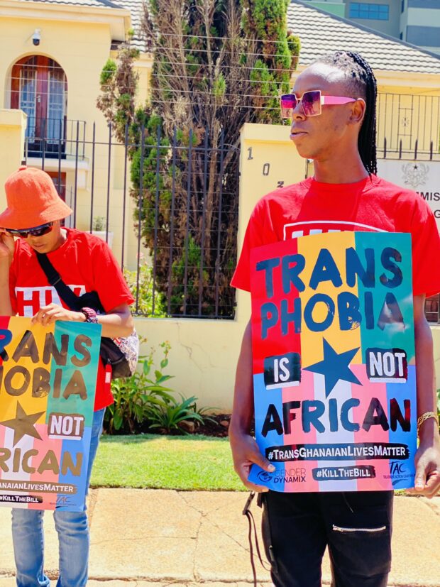 Two protesters in red shirts holding a sign that reads “Transphobia is not African,” outside a government building.