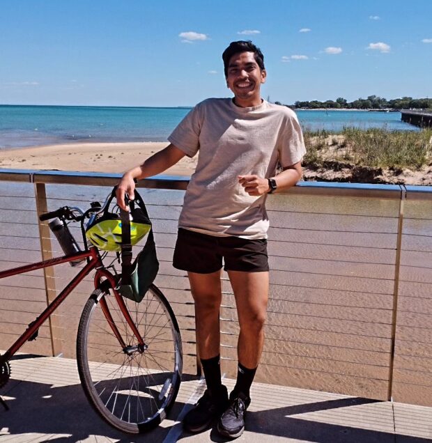 Person standing on a boardwalk beside a red bicycle, smiling with the ocean in the background.