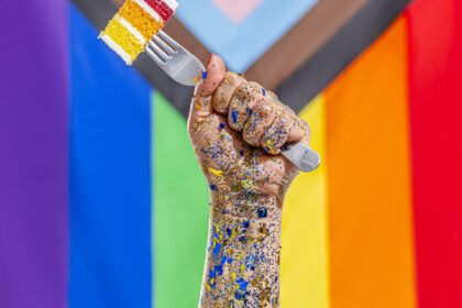 A painted hand holding rainbow cake on a fork in front of a Pride flag.