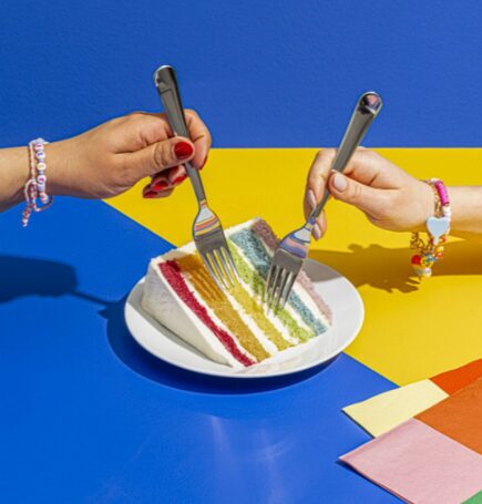 Two hands holding forks over a slice of rainbow cake on a blue and yellow background.