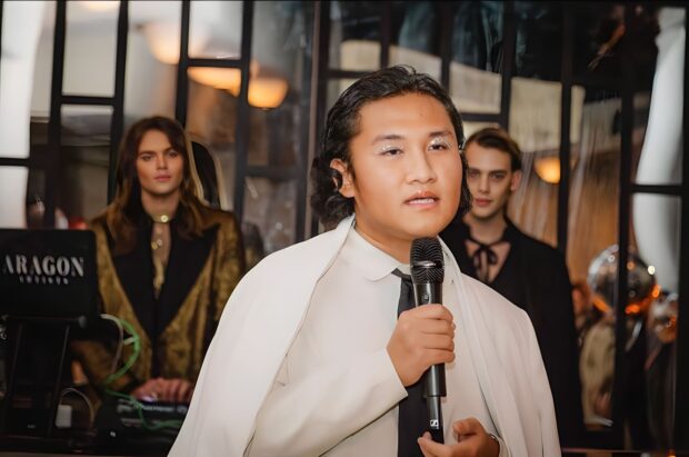 Person wearing white, speaking into microphone, with people in the background.