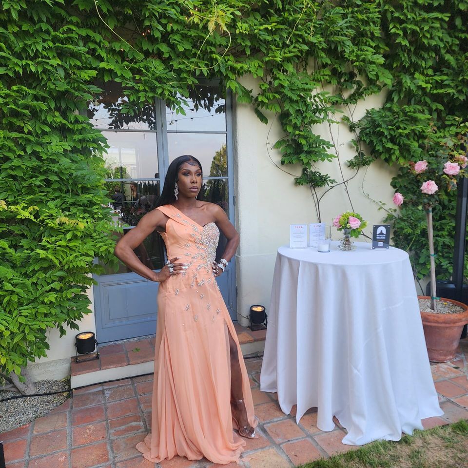Person in long coral gown in front of a glass door and beside a white table.