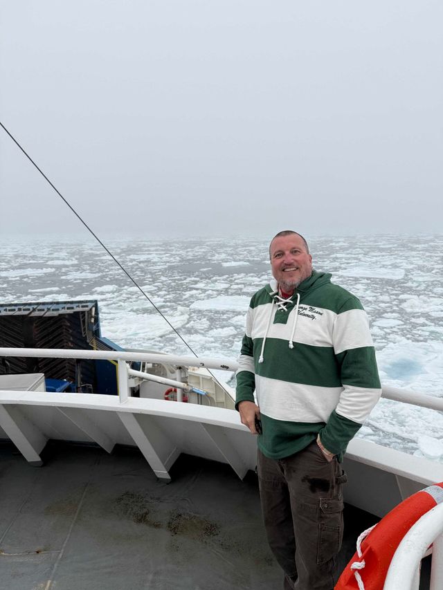 Person standing on a ship deck wearing a green and white striped sweater, smiling with icy ocean waters in the background.