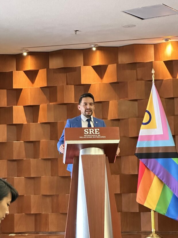 Person at a podium speaking beside a Pride flag in a wood-panelled room.