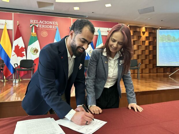 Two people signing documents at a table during an official event with national flags behind them.