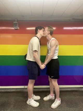 Couple kissing in front of a wall painted with rainbow stripes, holding hands and facing each other.