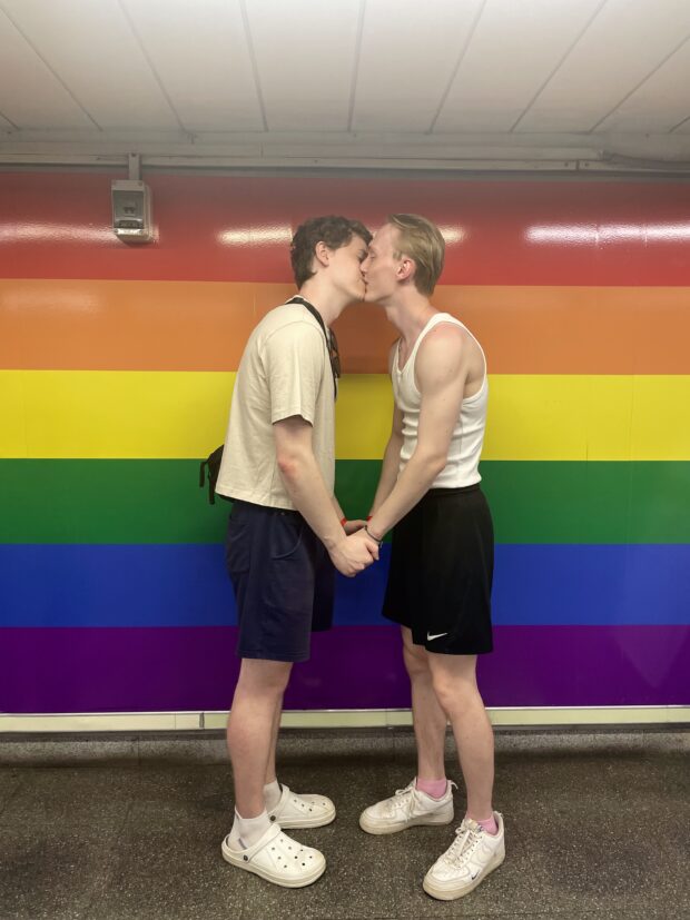 Couple kissing in front of a wall painted with rainbow stripes, holding hands and facing each other.