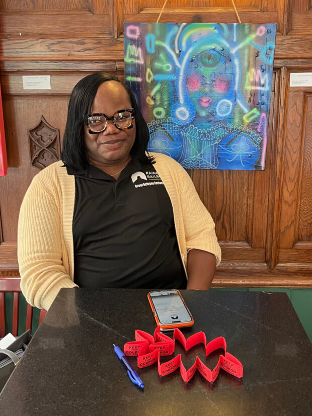 Person wearing glasses and a Rainbow Railroad shirt seated at a table with raffle tickets and a phone.