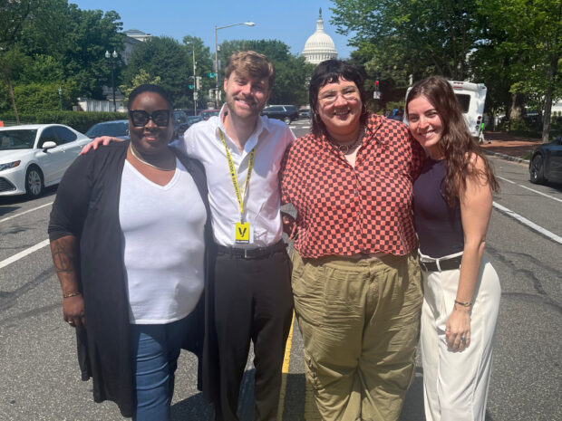 Four people standing together outdoors smiling, with the U.S. Capitol building visible in the background.