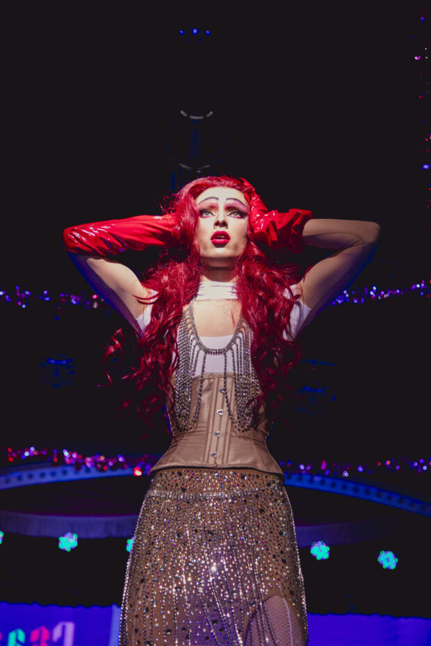 Drag performer with red hair and gloves posing onstage under bright lights.