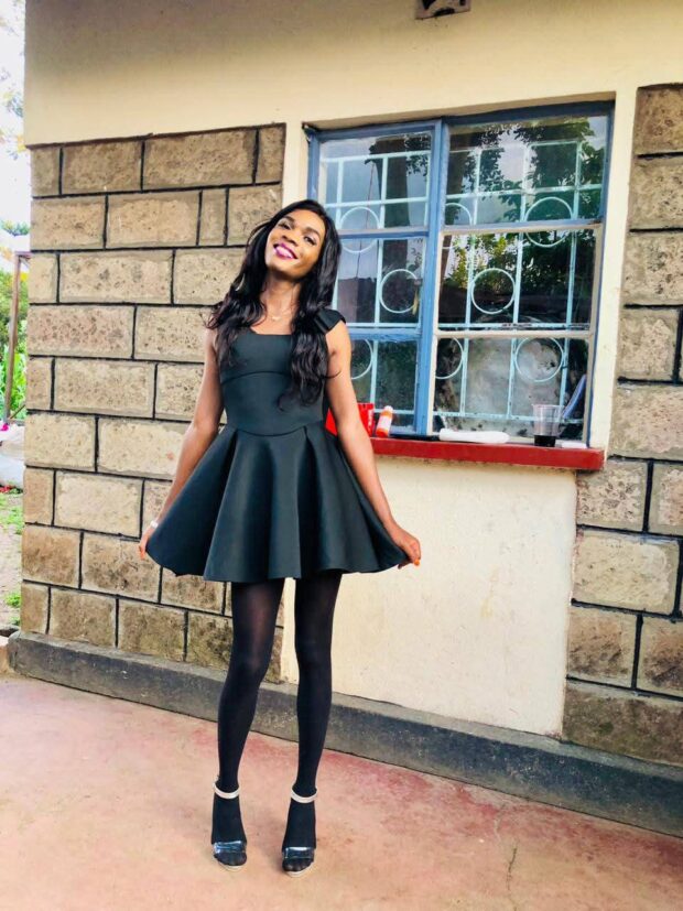 Person smiling in a sleeveless dark dress standing outside by a brick wall and window.