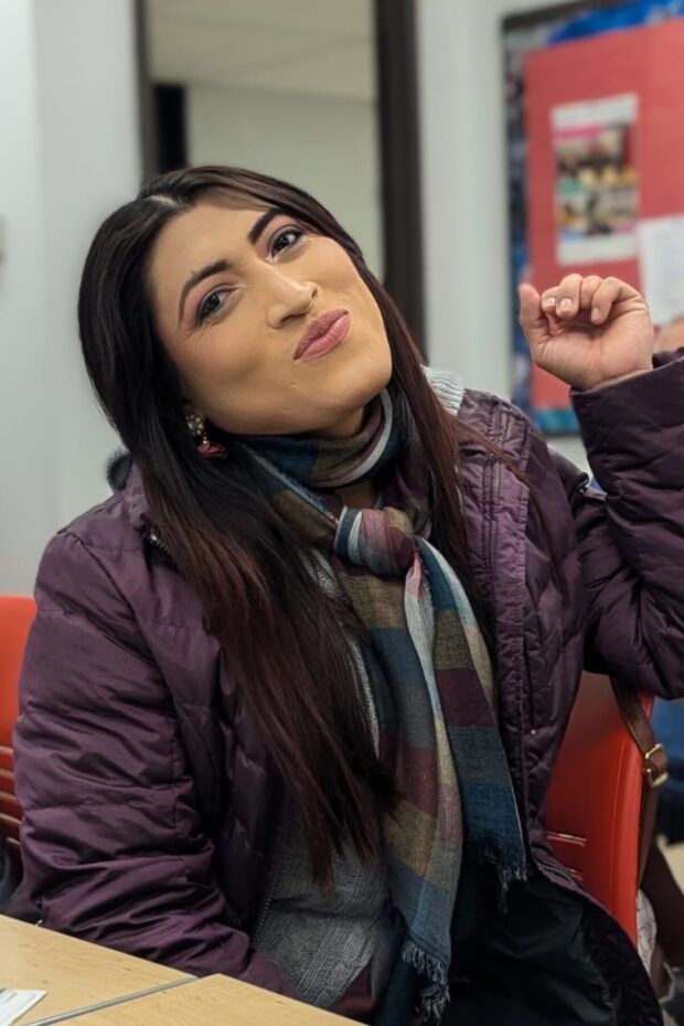 Person sitting indoors in a scarf and jacket, smiling.