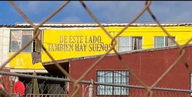 Building with yellow sign reading “DE ESTE LADO TAMBIÉN HAY SUEÑOS,” viewed through a chain-link fence.