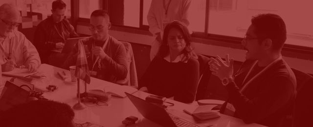 People seated at a table in discussion, with notebooks and laptops in front of them, under a red color overlay.