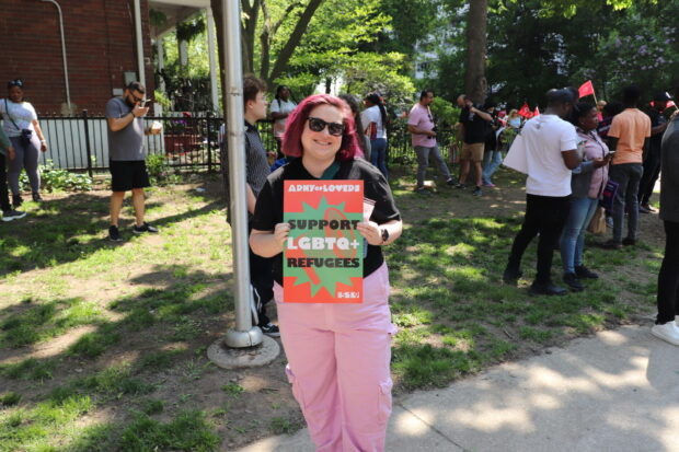 Person with pink hair holding a sign reading ‘Support LGBTQ+ Refugees’ outdoors with people in the background.