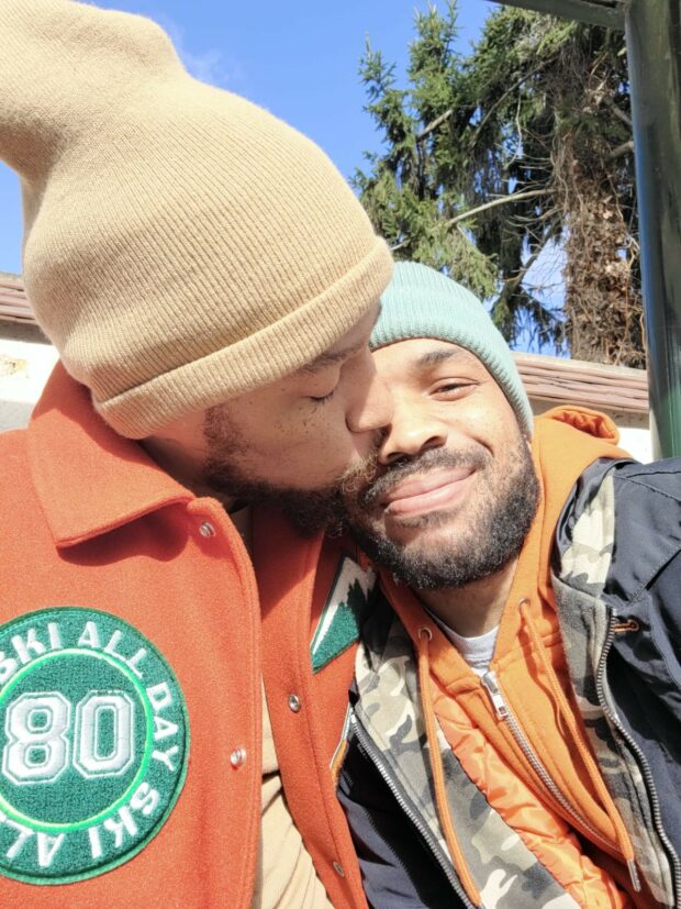 Two people in winter hats sharing a kiss and smiling outdoors.