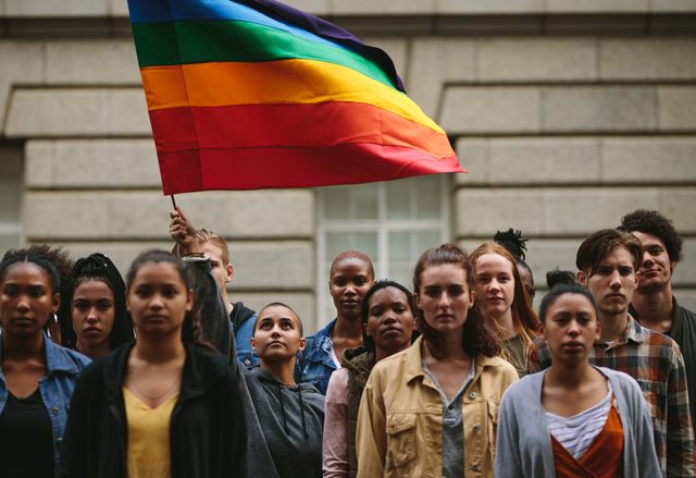 A group of people stand together as one person raises a rainbow Pride flag above them.