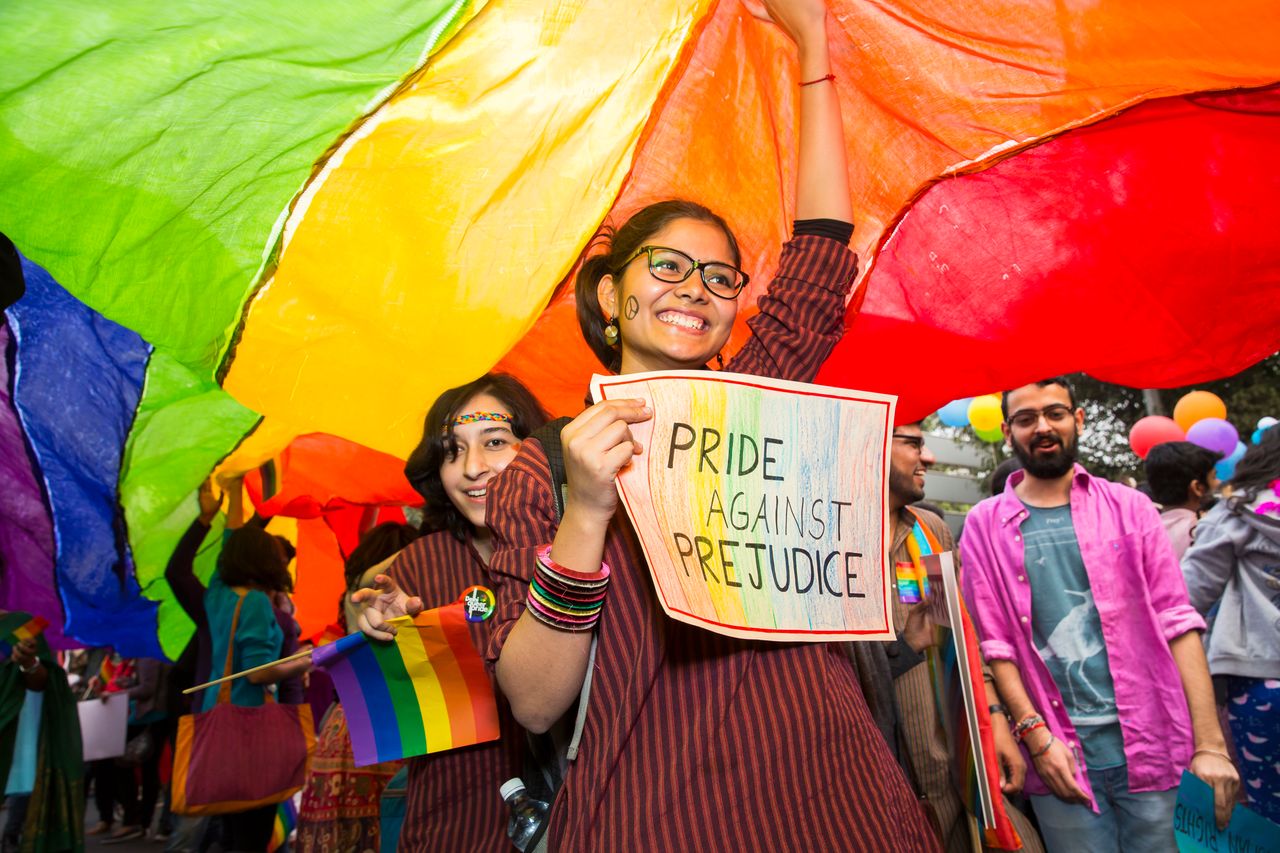 People at a Pride event standing underneath a large Pride flag holding up handheld Pride flags and signs 'Pride against injustice' while smiling.