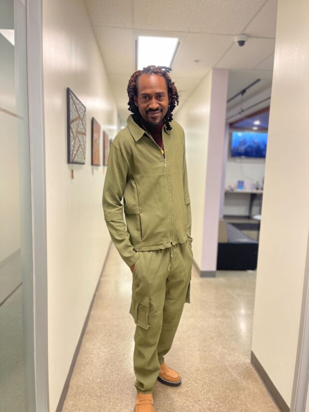 Person standing in a hallway wearing an olive-green outfit and smiling.